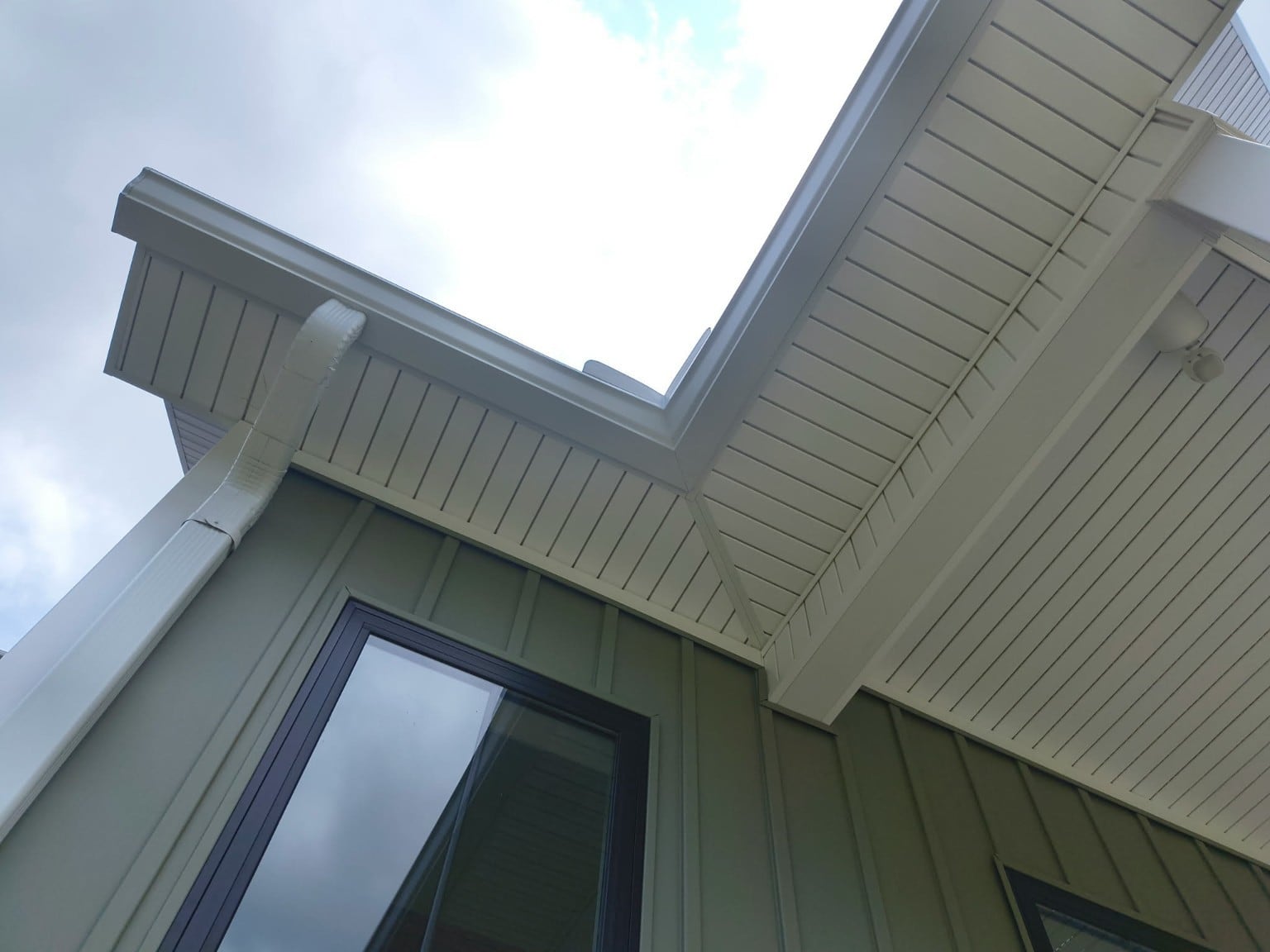 White gutter and downspout beneath a roof corner above green siding.